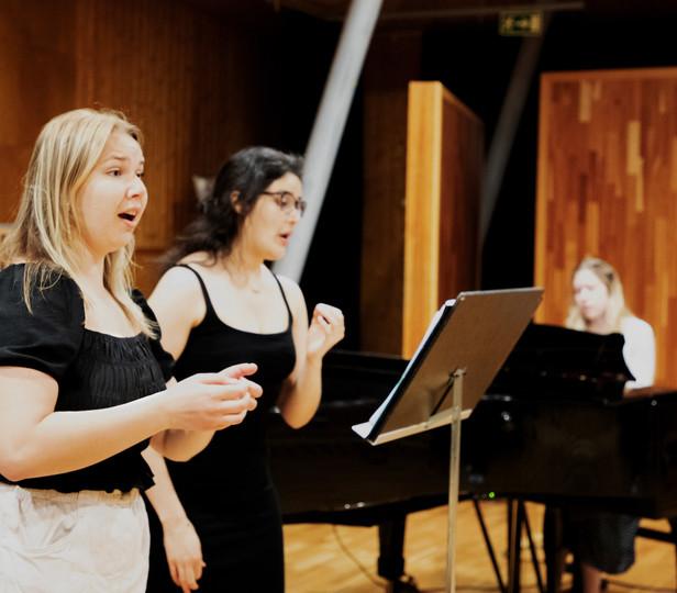 Two singers in a wood-paneled rehearsal studio, with a collaborative pianist at the keyboard, 在后台. Both singers are focused on sheet music on a stand in front of them